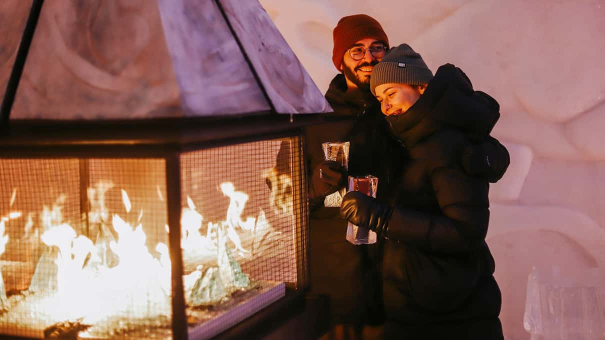 A couple in winter hats holding warm drinks beside a fire in a glass hearth at the Hôtel de Glace at night