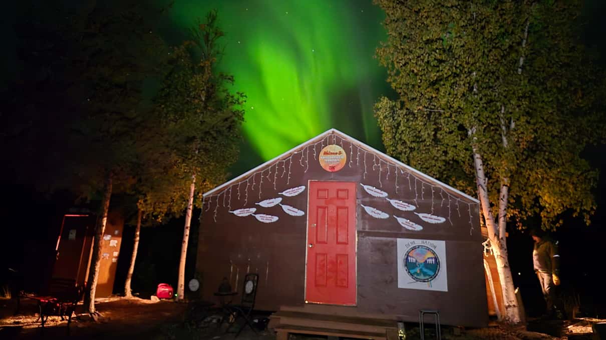 A wooden cabin with a lit front under a green aurora showing through the trees