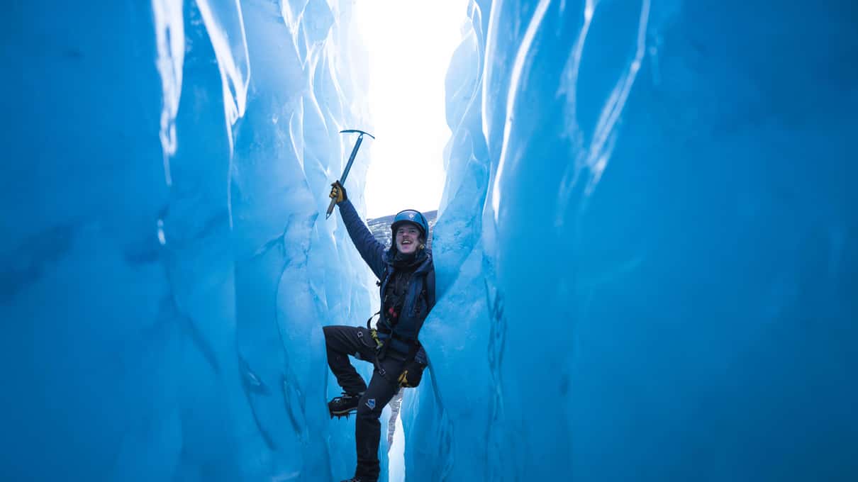A climber with an ice axe looking up from inside a deep blue moulin on the glacier