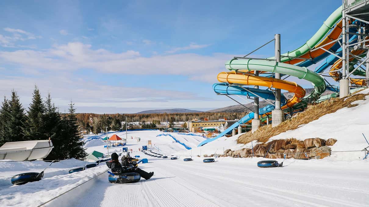 Riders sliding down a groomed snow tubing run at Valcartier, with pine trees and tall slides behind
