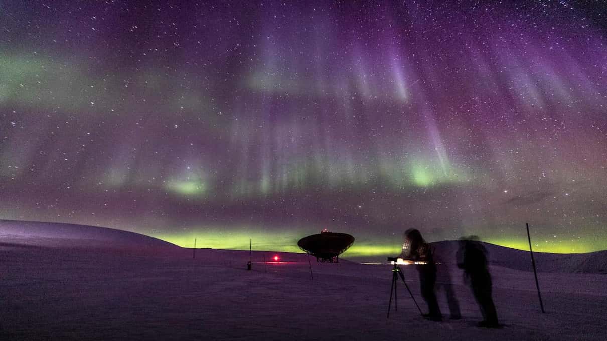 Fotógrafos con trípodes sobre la nieve bajo una aurora violeta y verde