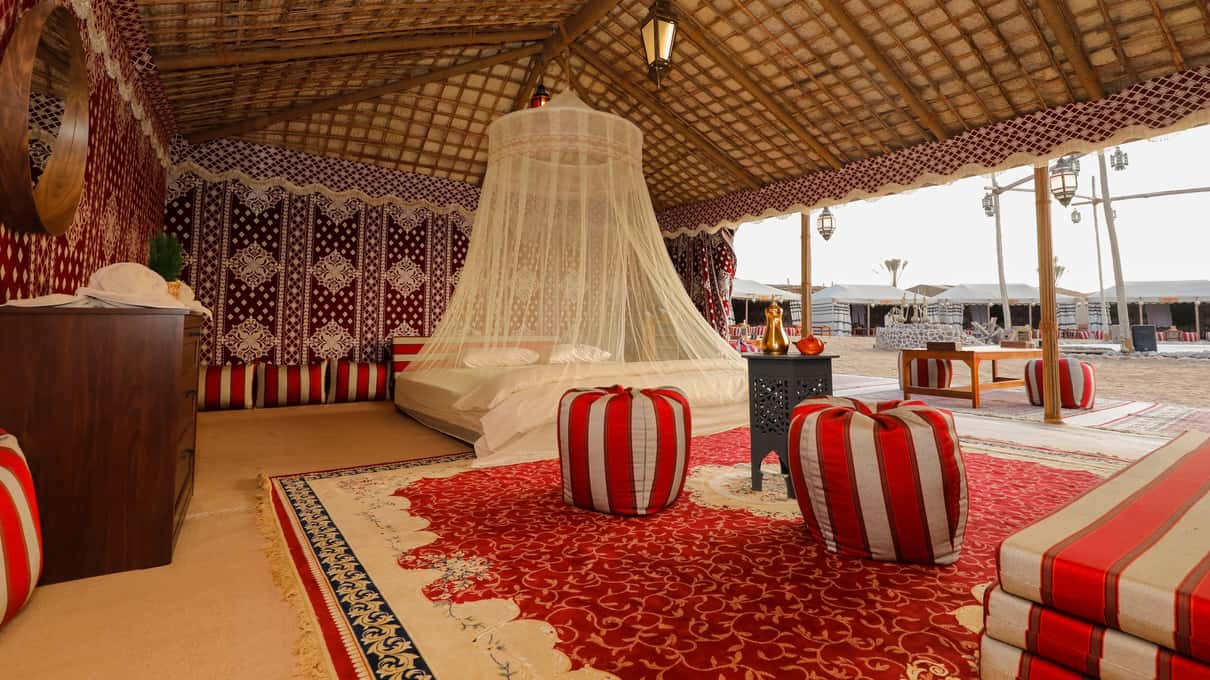 Interior de una carpa del campamento con cama bajo mosquitera, alfombras rojas y pufs de rayas