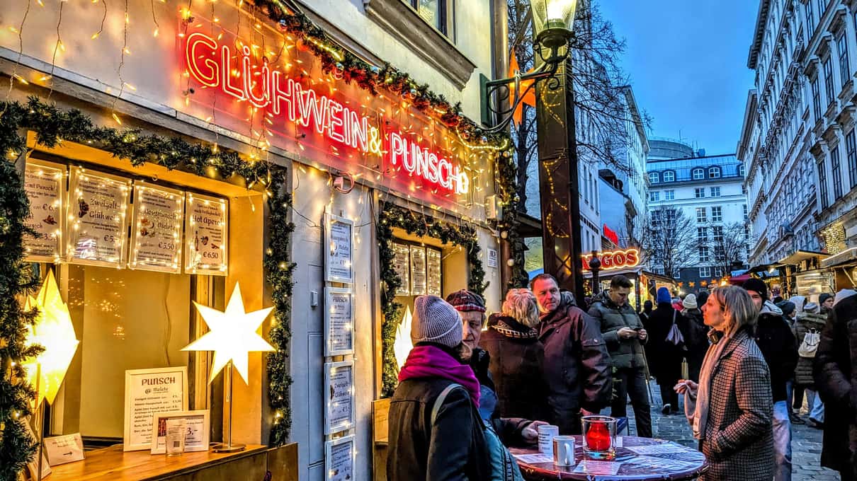 A Glühwein and Punsch stand with a red neon sign and paper stars, with people holding mugs in the street at blue hour