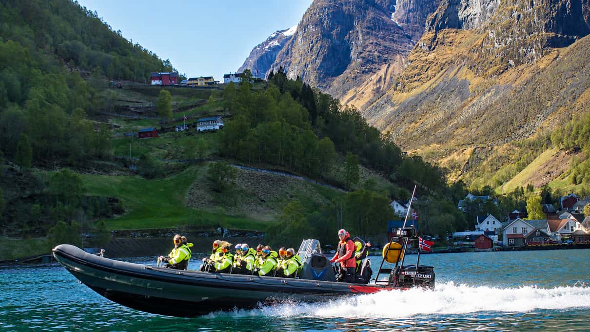 A RIB boat full of passengers in orange thermal suits idling beside the steep green shore of the Nærøyfjord