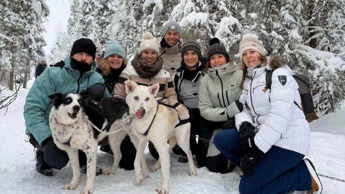 A smiling group of guests in winter jackets crouching with two huskies in a snowy forest