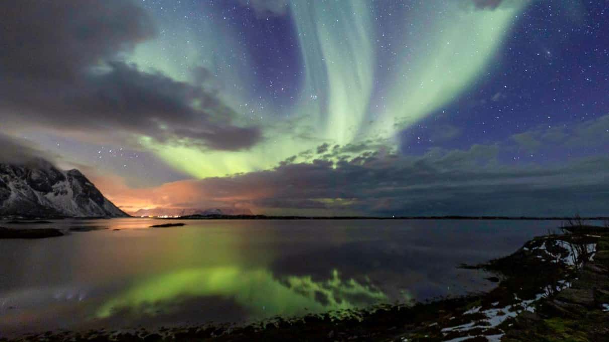 A green aurora breaking through a gap in the clouds above jagged Lofoten peaks and calm reflecting water