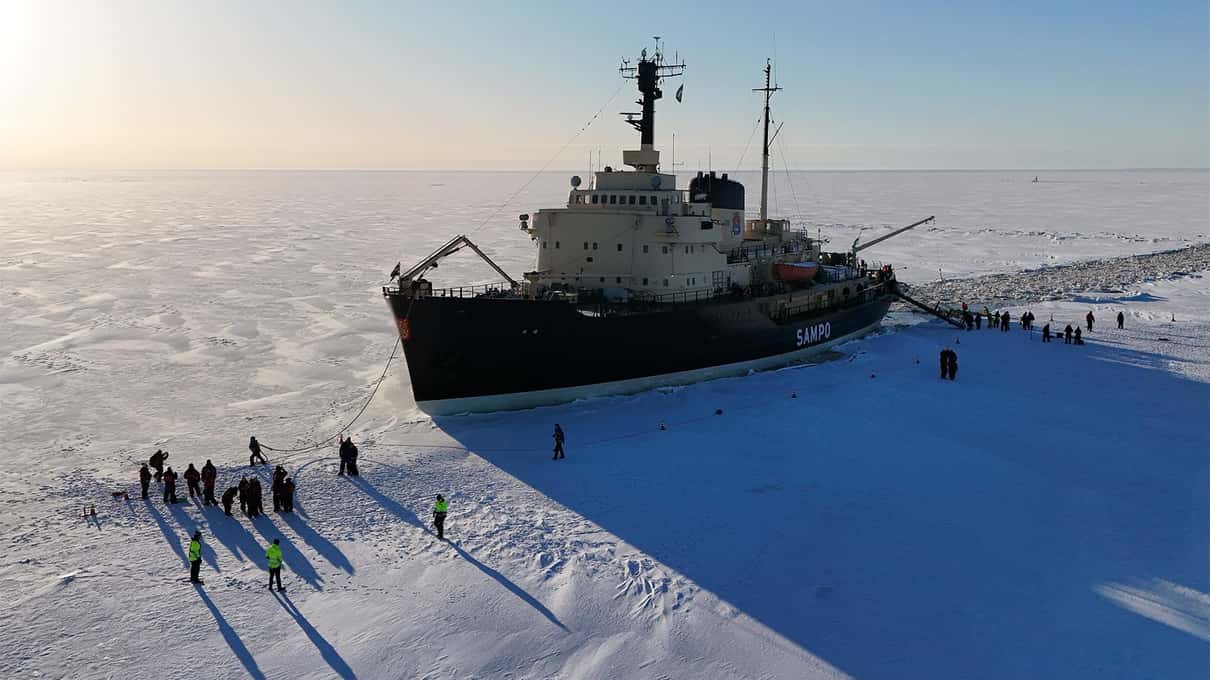 The black hull of the icebreaker Sampo stopped in white sea ice, with passengers walking on the frozen sea beside it