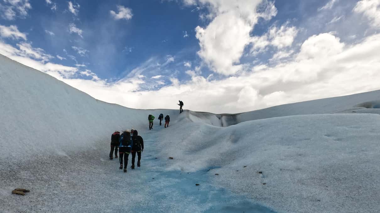 A group of trekkers climbing the glacier beside a blue meltwater channel under broken cloud and sun