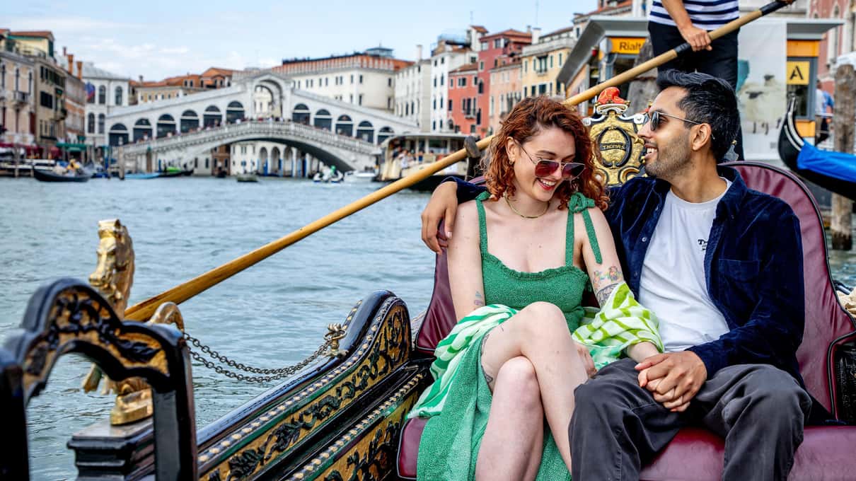 Couple rieur assis dans une gondole sur le Grand Canal, derrière eux le pont du Rialto et la rame du gondolier