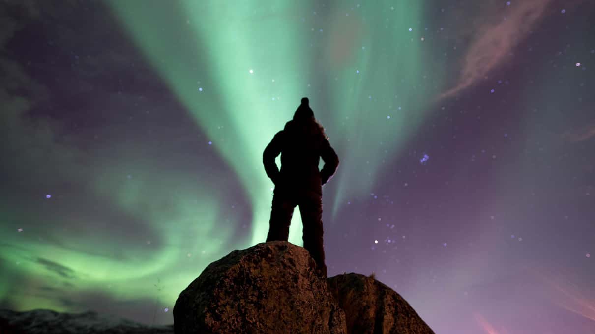 Silhouette debout sur un rocher, de nuit, sous une aurore boréale verte et violette