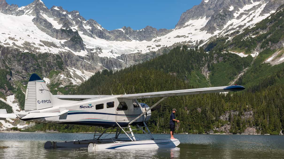 Floatplane resting on a turquoise alpine lake below snowy peaks with a passenger standing on the float