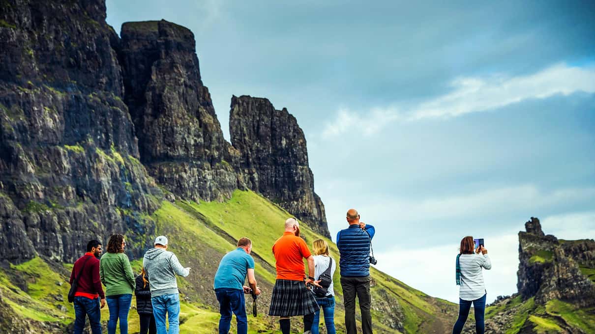 Tour group including a guide in a kilt looking up at the dark rock pinnacles and green slopes of the Quiraing