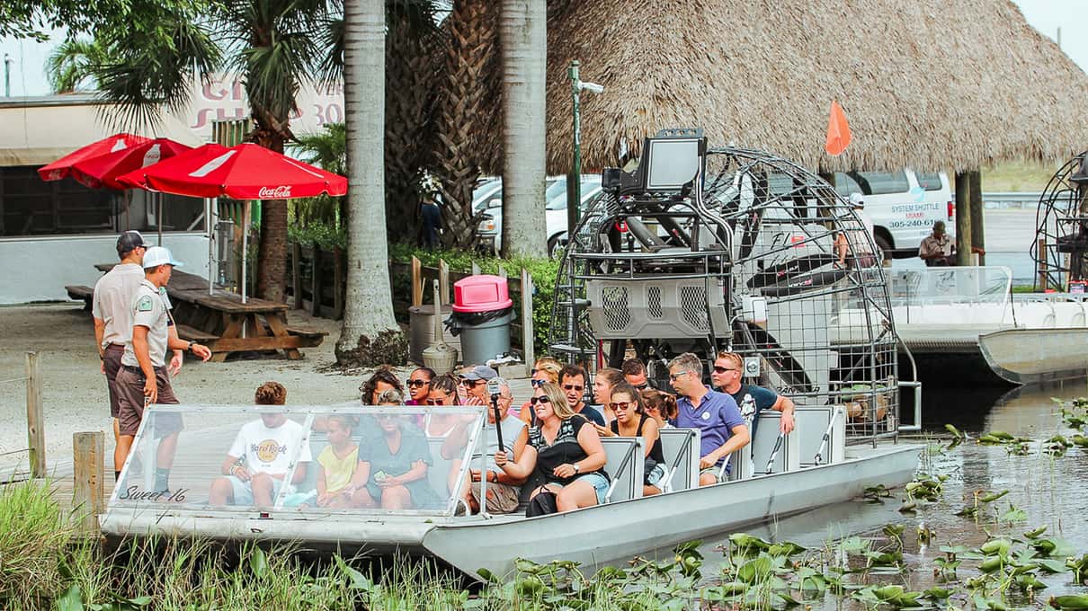An airboat loaded with guests waiting at Gator Park's dock next to a thatched chickee and red umbrellas