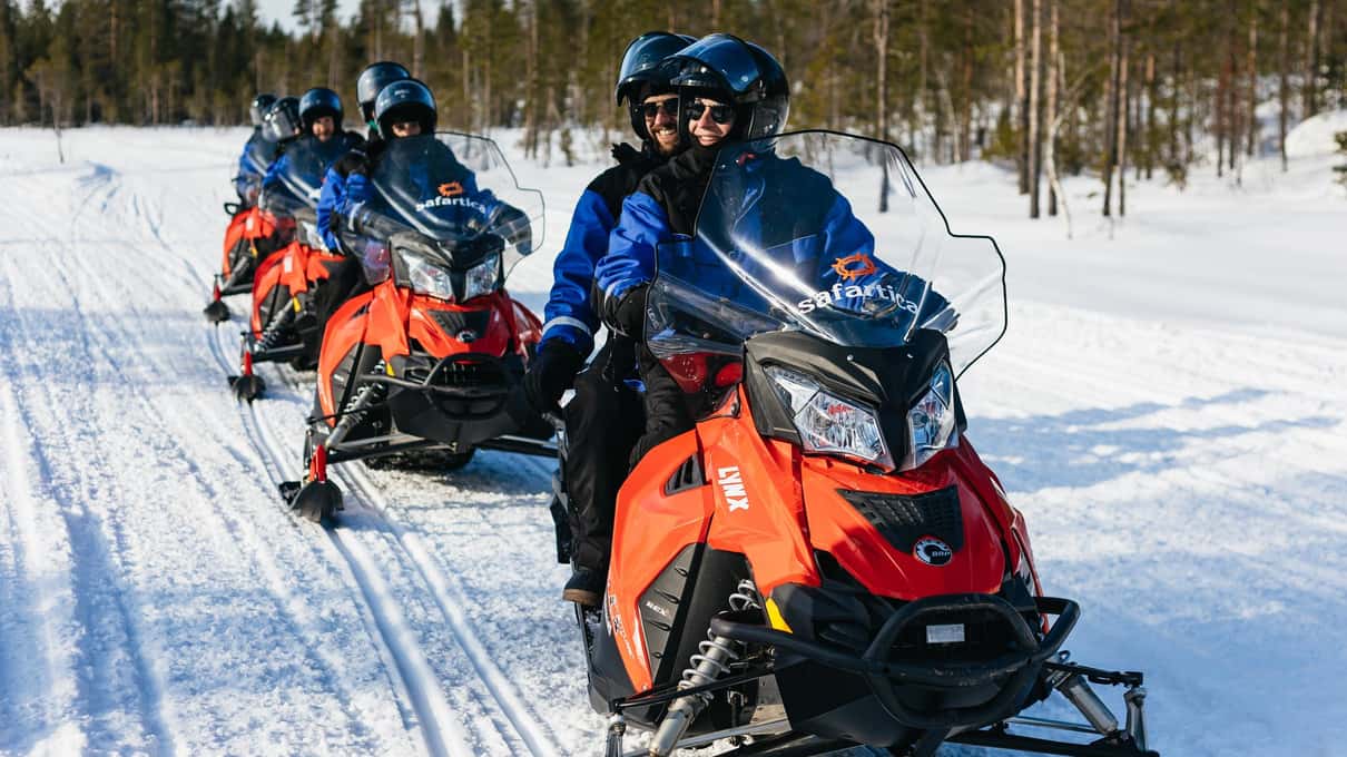 A line of red snowmobiles with riders in blue overalls crossing a snowy pine forest near Rovaniemi
