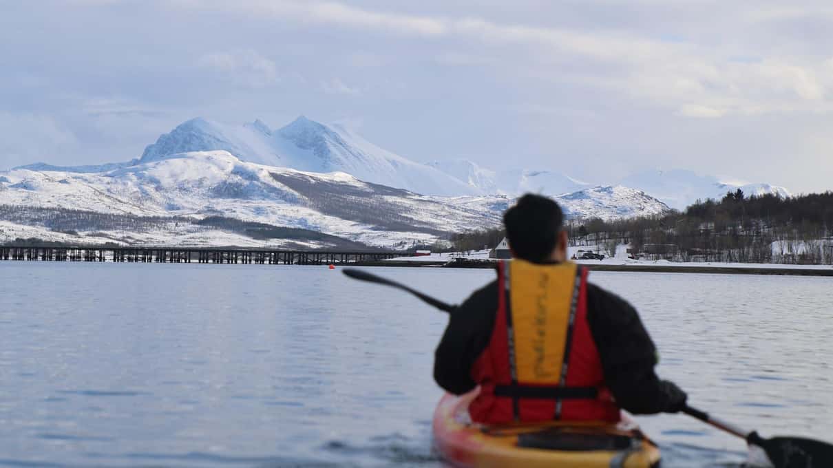 A paddler in a yellow buoyancy vest looking across the fjord at a snow-capped peak and a long bridge