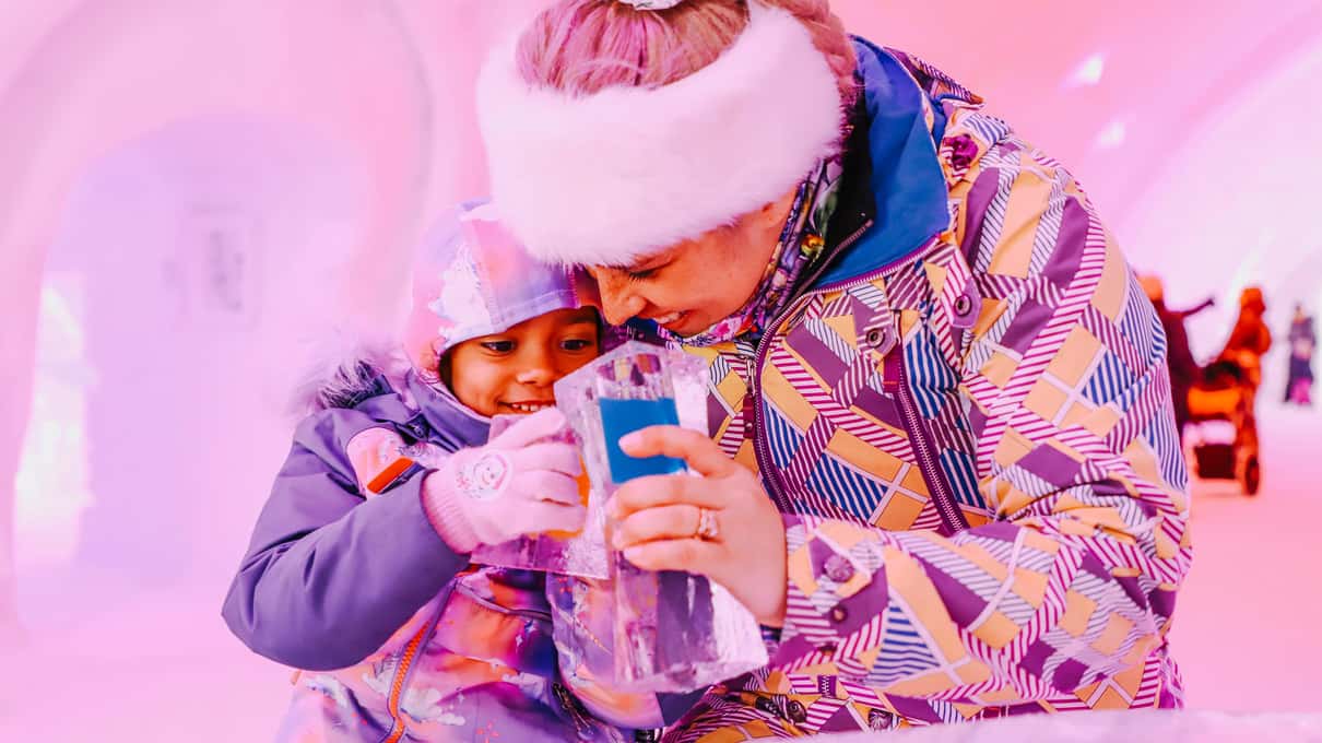 A mother and a small child in snowsuits standing at the ice bar inside the Hôtel de Glace
