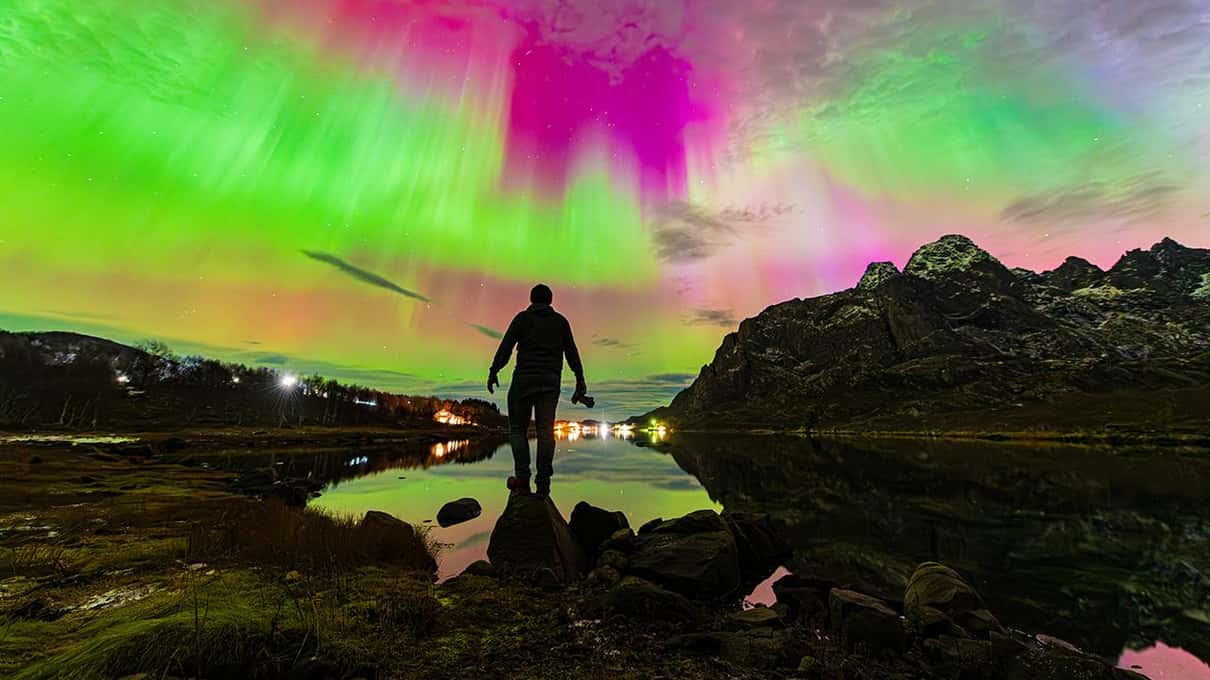 A man standing on rocks by a still lake under a green and pink aurora, with Lofoten mountains reflected in the water