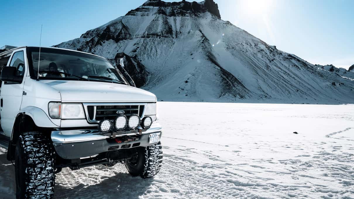 En hvid Super Jeep med kæmpe dæk parkeret på sne under en mørk tinde med striber af sne