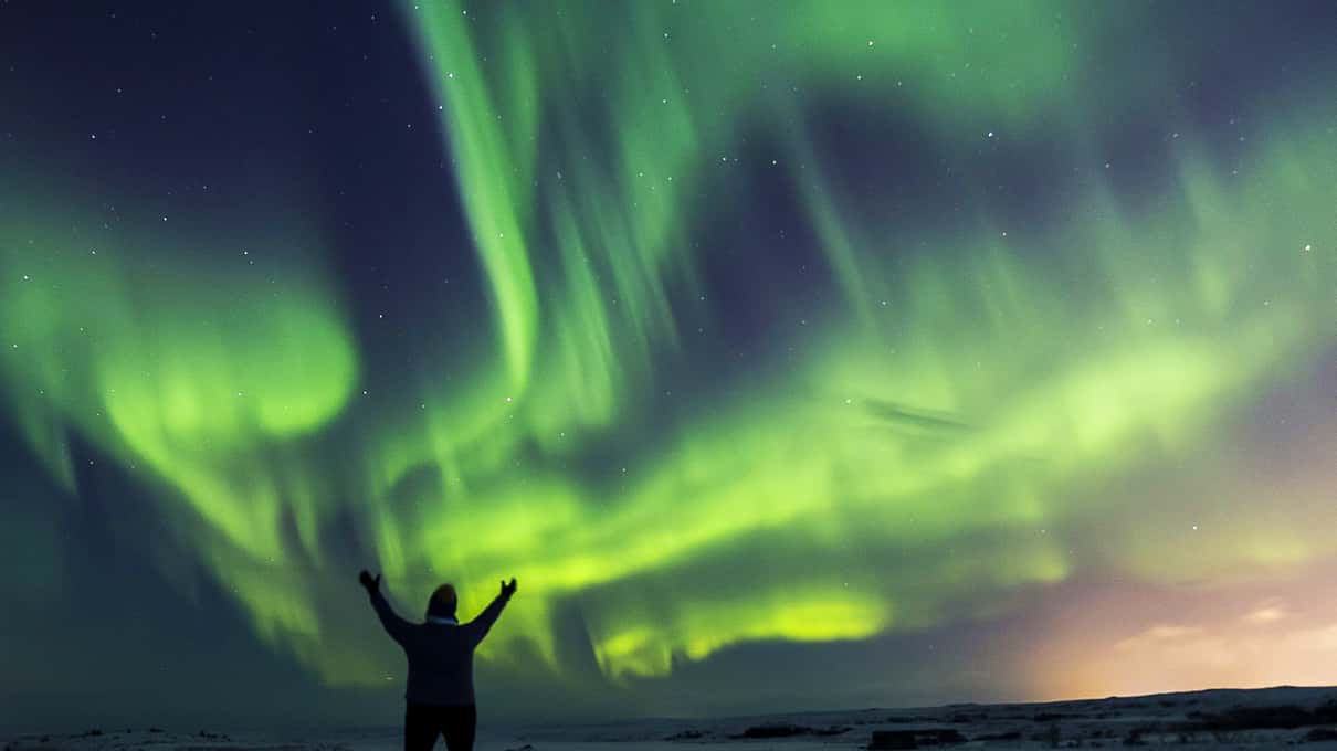 Reisender mit erhobenen Armen unter einem weiten grünen Nordlichtbogen in Island