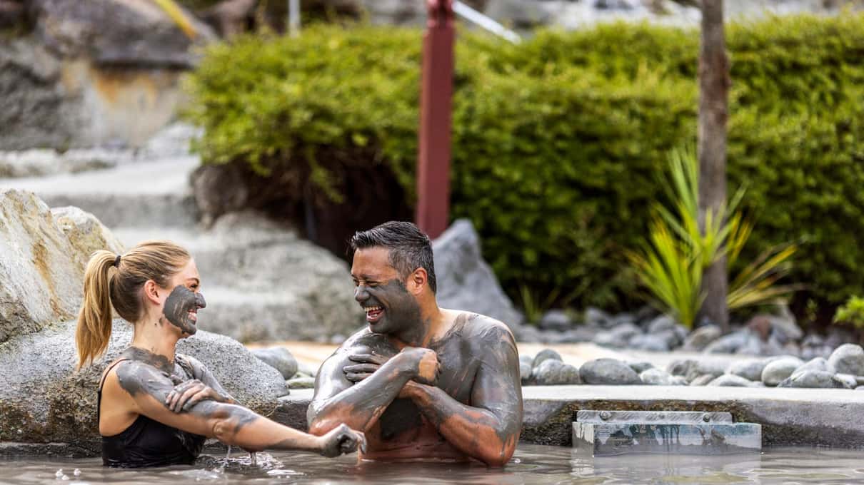 A laughing couple with grey geothermal mud smeared on their faces at Hell's Gate