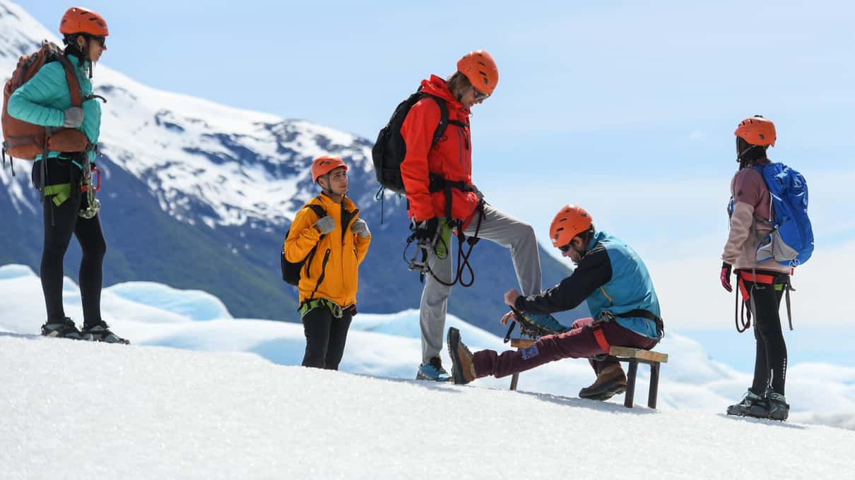 A guide kneeling on the glacier to fit crampons while hikers in helmets and harnesses wait their turn