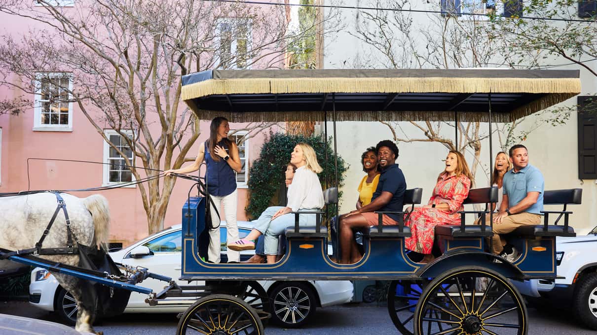 Smiling riders on the benches of a blue Palmetto carriage beside a pastel house and spring blossom
