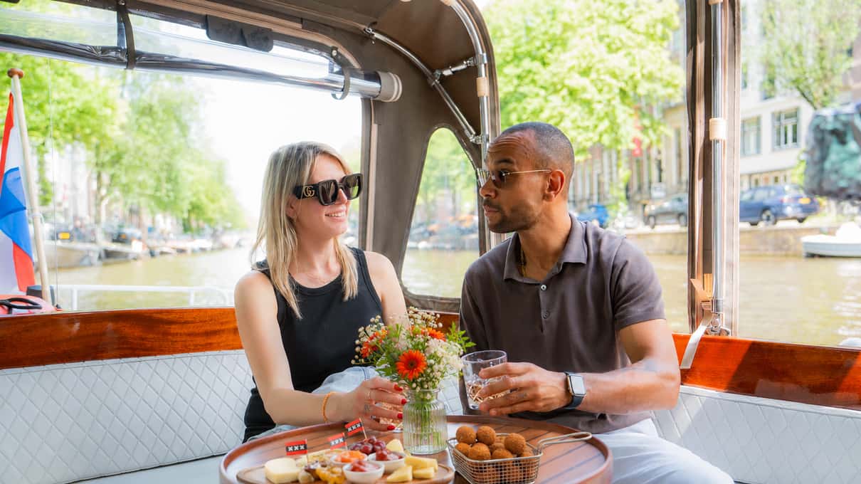 Twee passagiers met drankjes en een schaal bitterballen aan boord van een open sloep op de gracht