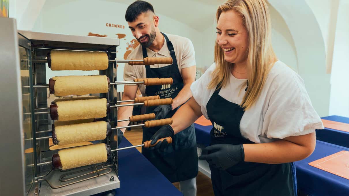 A baker and a smiling guest turning trdelník dough on wooden spits over a hot grill