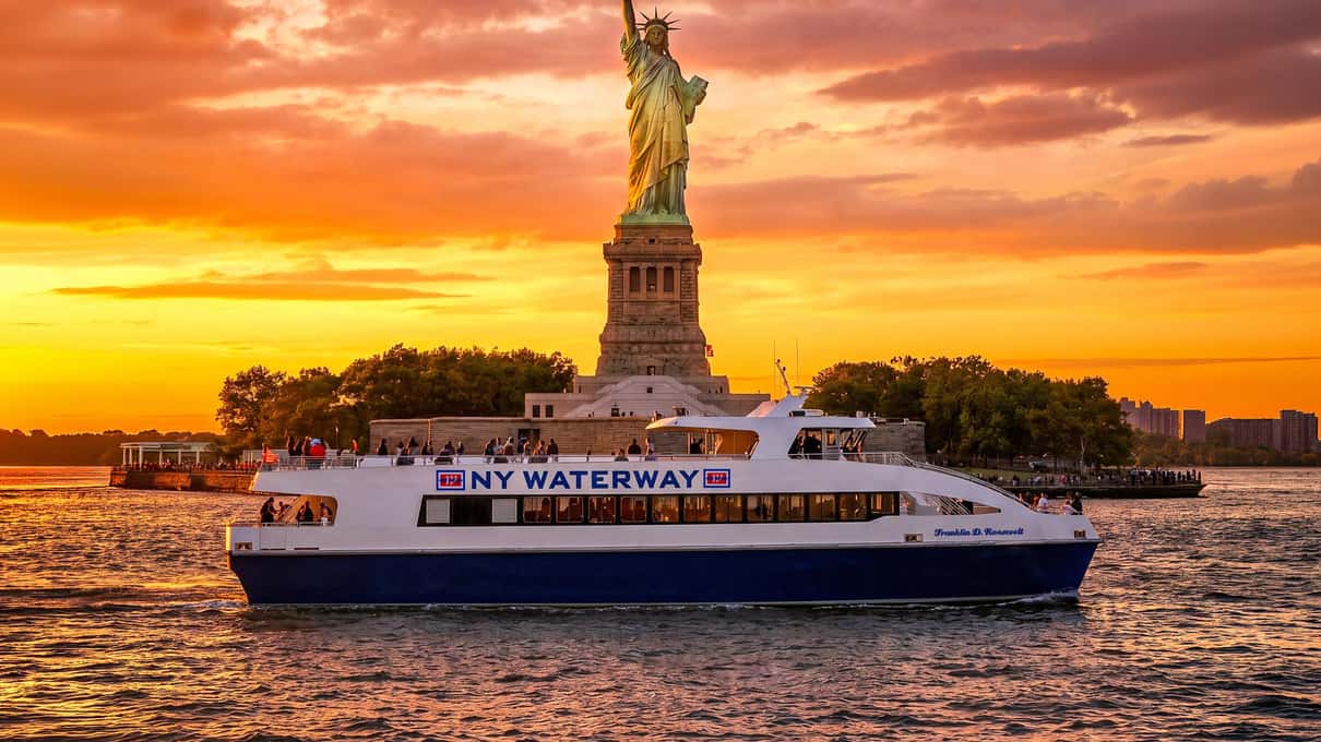 NY Waterway sightseeing boat passing the Statue of Liberty under an orange sunset sky