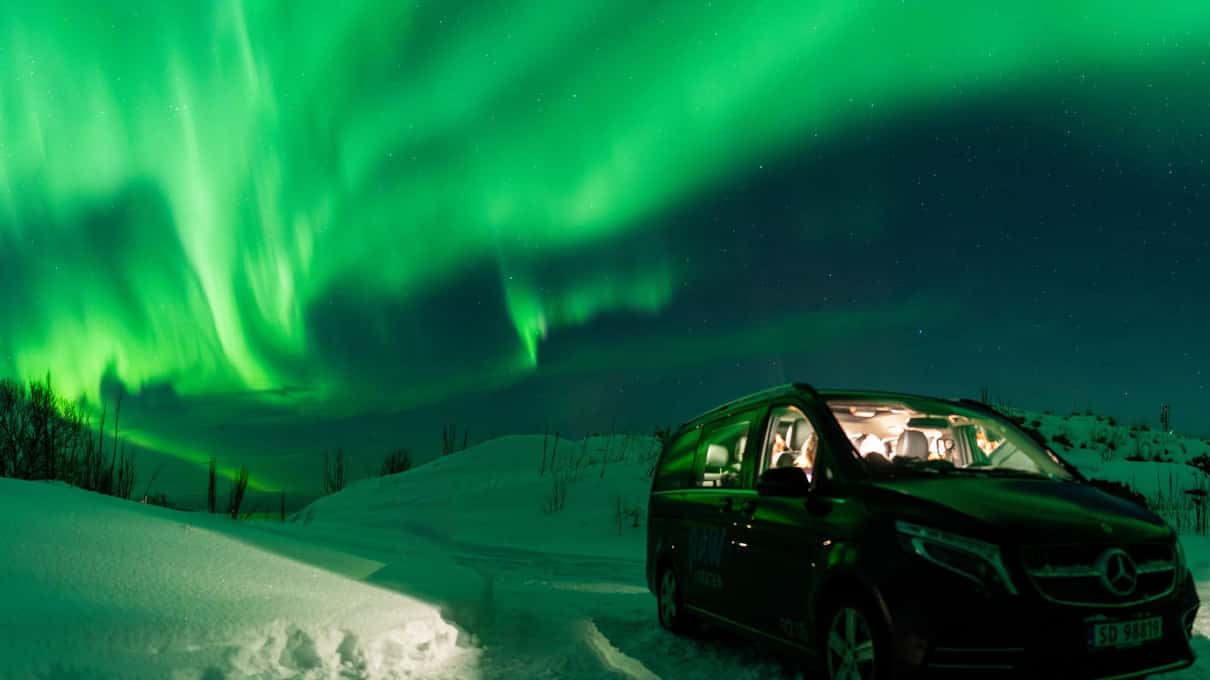 A black Mercedes van parked in deep snow under a bright green aurora in Lofoten