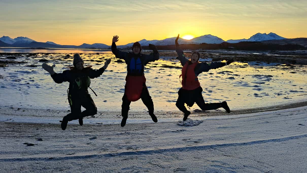Three paddlers in drysuits jumping on a snowy beach at sunset after a winter kayak tour