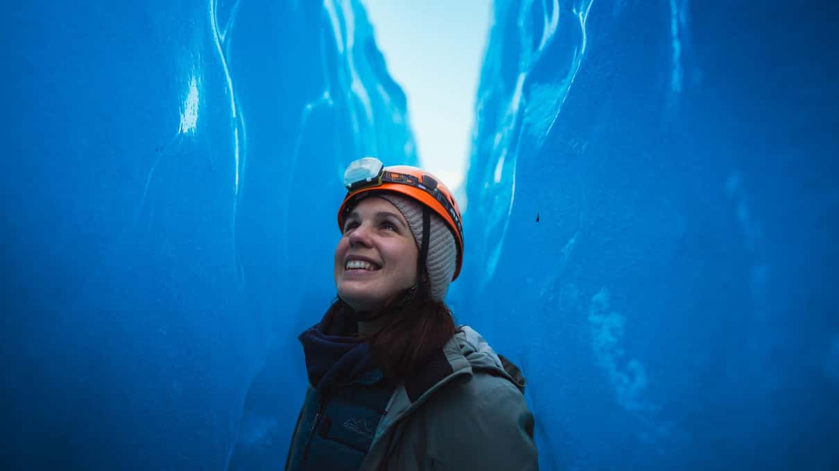 Smiling hiker in an orange helmet looking up between towering walls of deep blue glacier ice on Vatnajökull
