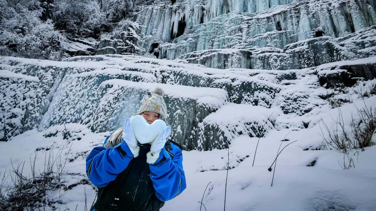 A hiker in blue thermal overalls standing in deep snow below the frozen waterfalls of Korouoma canyon