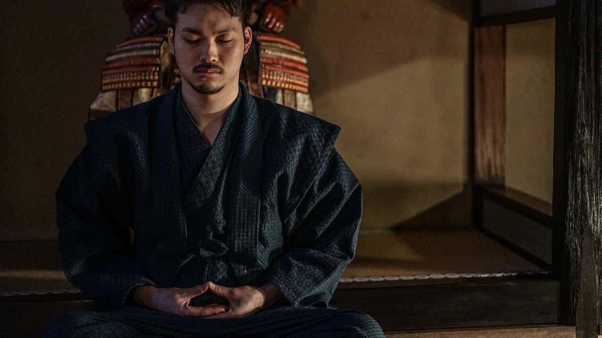 Ein Mann sitzt in Zazen-Meditation auf Tatami vor einer ausgestellten Samurai-Rüstung