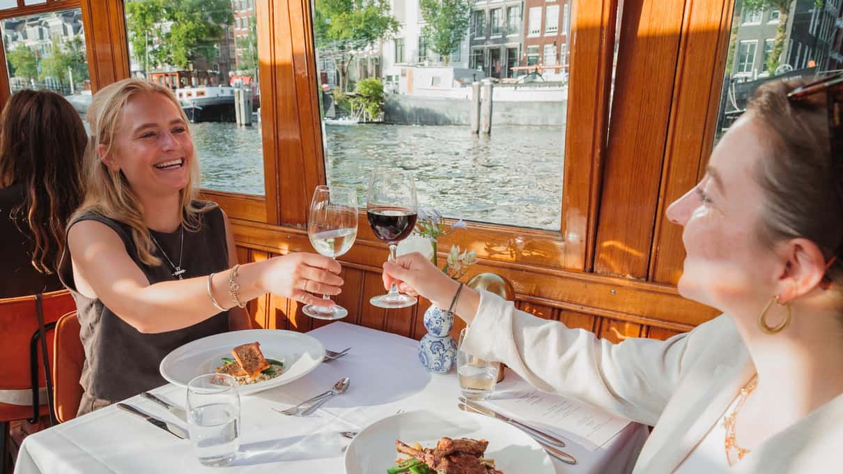 Twee gasten proosten aan een gedekte tafel bij het raam van een salonboot tijdens een dinerrondvaart