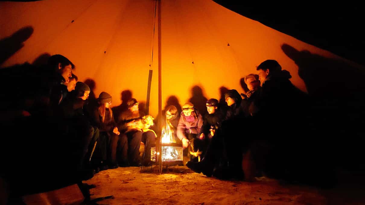 Guests in thick winter clothes sitting around a wood fire inside a lávvu tent at night