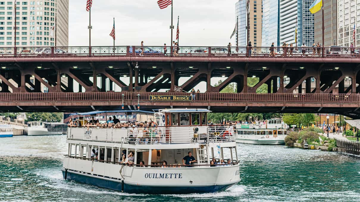 A white Wendella cruise boat with passengers on the top deck passing under a steel bridge on the Chicago River