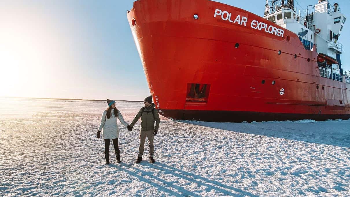 Two visitors walking on the sea ice beside the red bow of the Polar Explorer icebreaker