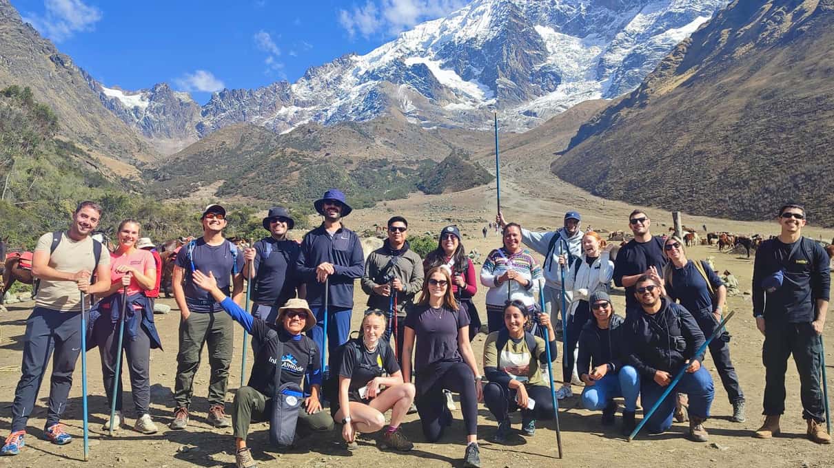 A large group of hikers jumping in the air on a grassy slope below snow-covered mountains