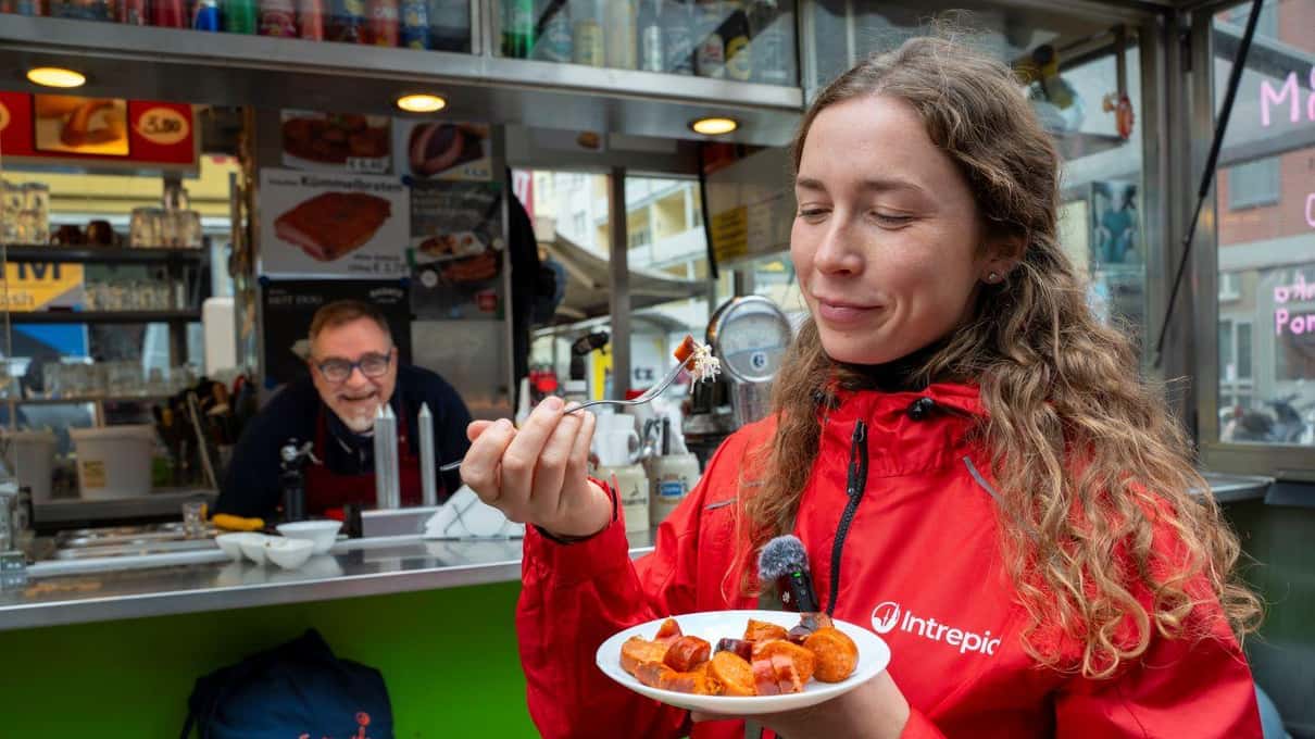 A smiling guide in a red jacket tasting a plate of goulash at a Viennese sausage stand