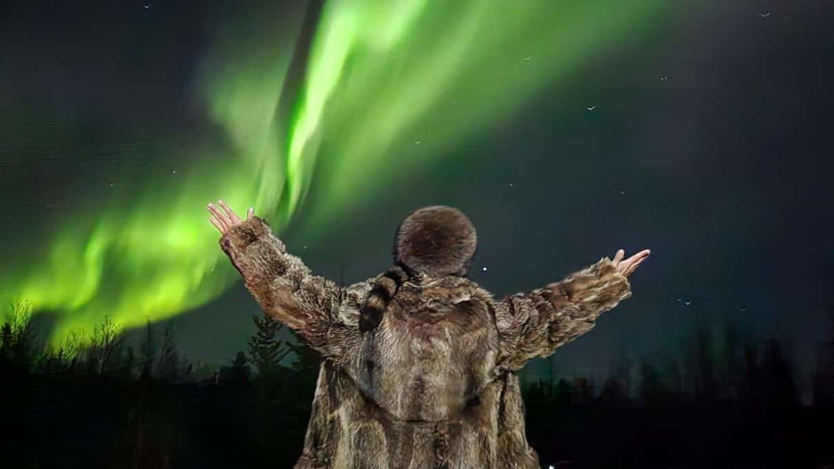 A guest in a fur-trimmed coat with arms spread wide under a green aurora near Yellowknife
