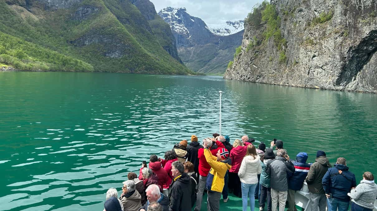 Passengers lining the rail of a fjord ferry as it sails green water toward snow-streaked peaks