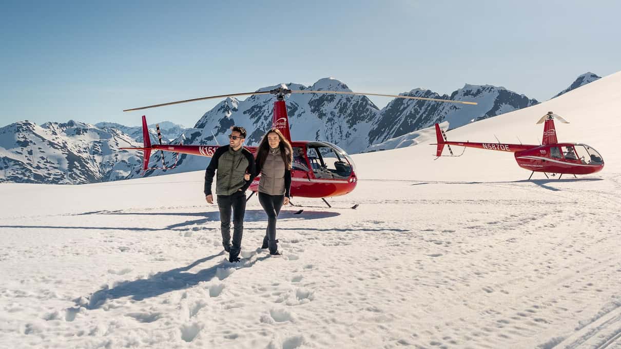 Two passengers walking across bright snow away from a pair of red helicopters parked on a glacier