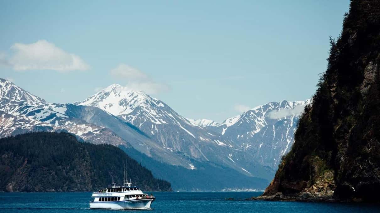 En lille hvid turbåd langt ude i en fjord under snedækkede tinder og en mørk klippevæg