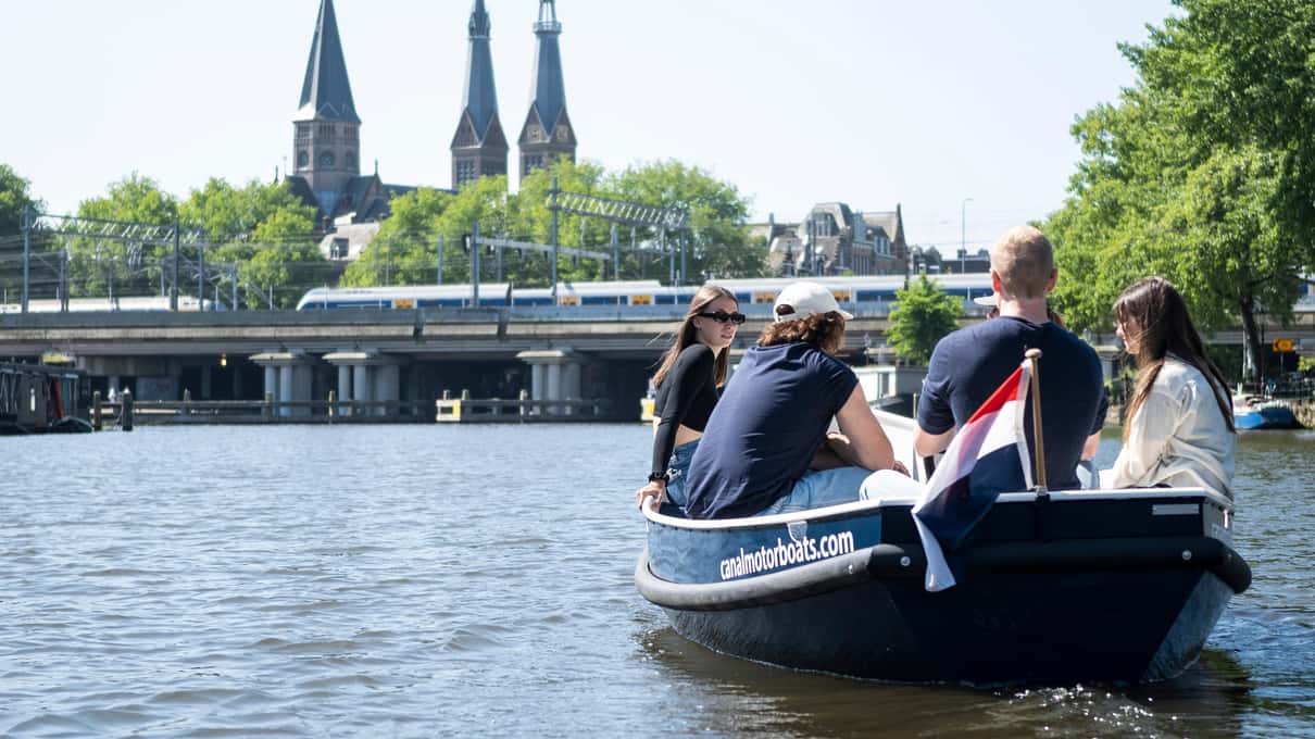 Passagiers in een kleine open motorsloep op een Amsterdamse gracht, met een brug en kerktoren op de achtergrond