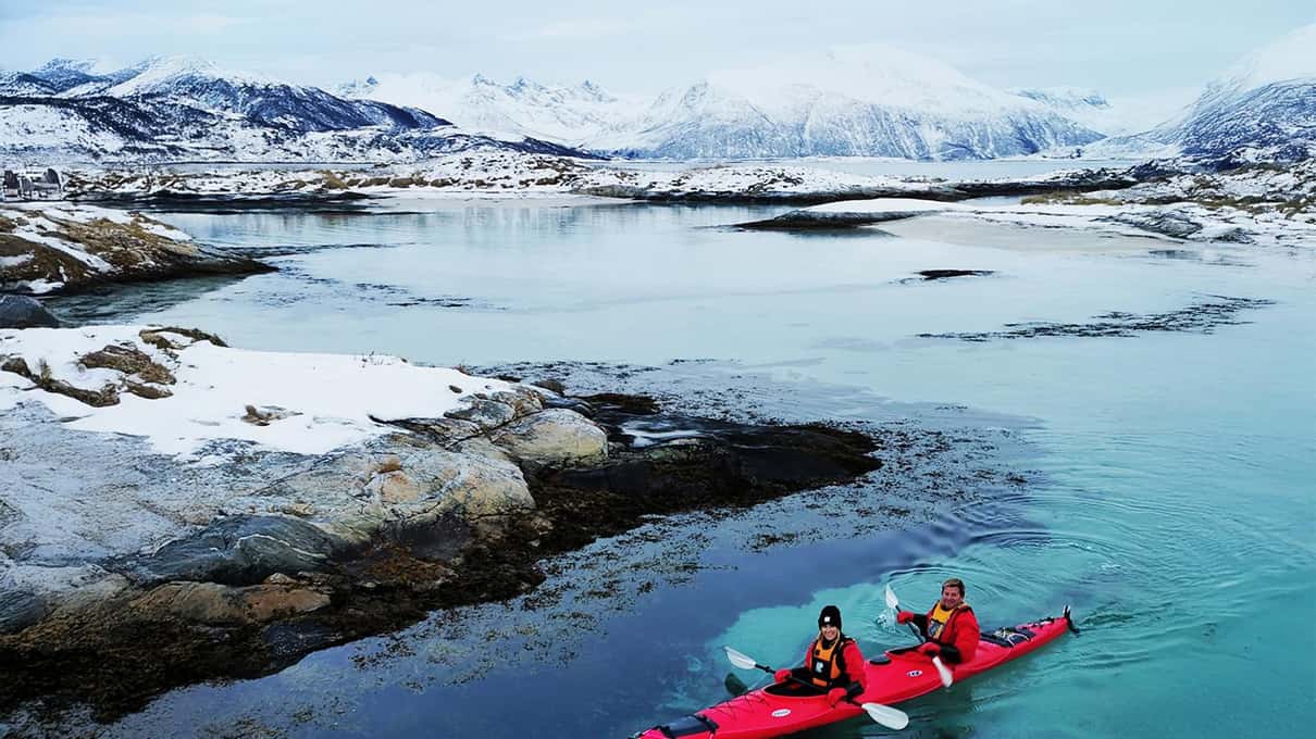 Two paddlers in a red double kayak in a turquoise channel between snowy rocks
