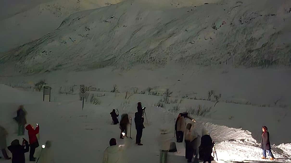 A tour group standing on a snowy slope at night under a faint green aurora near Alta