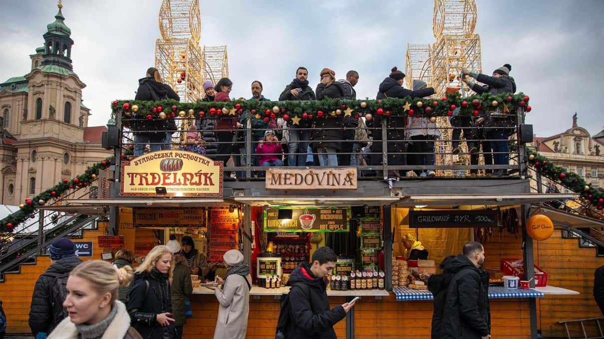 Visitors standing on the raised viewing platform above the wooden huts of the Old Town Square Christmas market, with the spires of the Týn Church behind