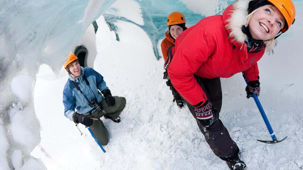 A guide in a red jacket helping a climber in a blue helmet at the edge of the ice
