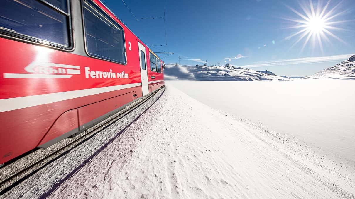 The side of a red Rhaetian Railway carriage running through deep snow under a blue sky